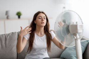 A woman sitting on a couch in front of a fan, looking desperate for cool air.