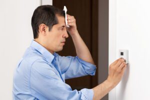 A man using a napkin to dry his forehead as he checks a thermostat.