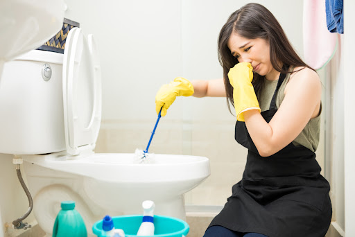 Woman holding her nose due to bad odors while plunging toilet in her bathroom with a bucket of cleaning supplies next to her.