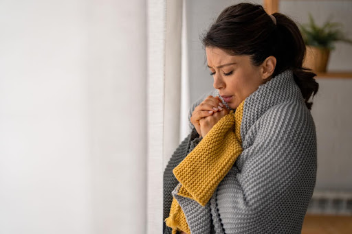 A woman blowing on her hands with a blanket wrapped around her in a home.