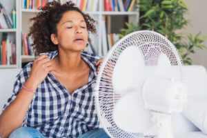 A woman in a warm house sitting in front of a fan to cool down.