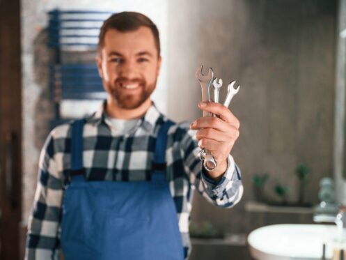 Smiling plumber holding a set of wrenches in a bathroom, ready for plumbing repair or maintenance.